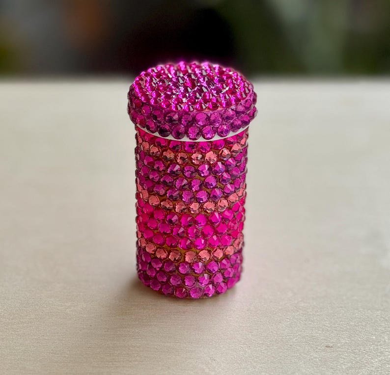 Op de afbeelding: Een roze en paarse juwelencontainer met een rond deksel. De container is bedekt met kleine, glinsterende strasssteentjes.