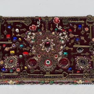 Op de afbeelding: Een rechthoekige donkerbordeauxrode doos versierd met talloze antieke koperen sleutels, kleurrijke gefacetteerde strass-steentjes en ingewikkelde metalen filigraanaccenten. Het oppervlak is dicht bedekt met deze decoratieve elementen.
