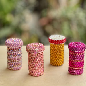 Op de afbeelding: Vier kleine, cilindrische containers versierd met kleurrijke strass steentjes. De containers zijn in tinten roze, oranje en geel.