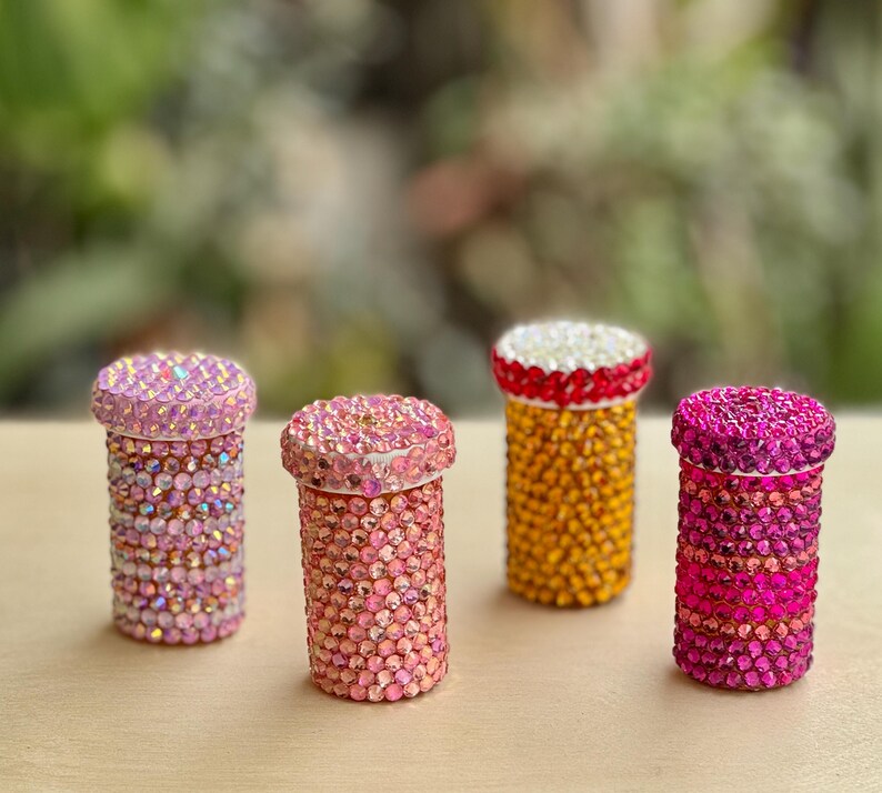 Op de afbeelding: Vier kleine, cilindrische containers versierd met kleurrijke strass steentjes. De containers zijn in tinten roze, oranje en geel.