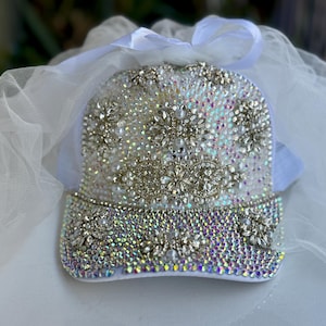 May include: A white baseball cap fully embellished with iridescent rhinestones and silver-toned floral accents. The brim and crown are covered in sparkling crystals. A white ribbon and tulle veil are attached to the cap, creating a glamorous accessory.