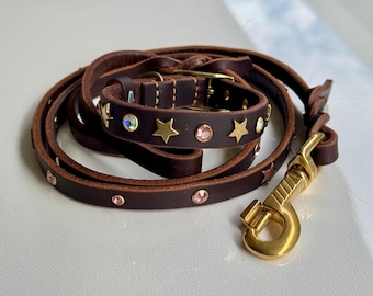 May include: A dark brown leather pet collar and matching leash set featuring gold-tone metal star-shaped studs and circular pink and iridescent crystal embellishments. The leash includes a gold-tone metal swivel snap hook for secure attachment.