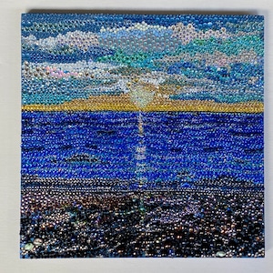 May include: A close-up of a beaded artwork depicting a seascape with a blue sky, white clouds, a yellow sandy beach, and a blue ocean. The ocean is made of blue beads, the beach is made of yellow beads, and the sky is made of blue, white, and iridescent beads.