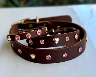 Puede incluir: Un conjunto de collar y correa de cuero marrón oscuro con herrajes dorados. El cuero está adornado con tachuelas circulares de cristal rosa y detalles dorados en forma de corazón dispuestos en un patrón repetitivo a lo largo de las correas.