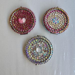 Op de afbeelding: Drie ronde, zilverkleurige compacte spiegels, elk bezet met iriserende strass. Eén heeft een roze hart, de andere hebben heldere, iriserende centra. Een decoratief accessoire.