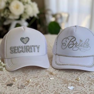 May include: Two white trucker hats. One hat has the word "SECURITY" spelled out in rhinestones with a rhinestone heart above. The other hat has the word "Bride" spelled out in rhinestones.