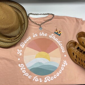 May include: A peach-colored t-shirt with the text "A Dove in the Shadows, Hope for Recovery" is paired with a straw hat, silver necklace, earrings, and brown sandals. The shirt features a sunset graphic.