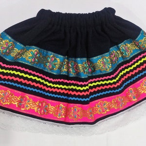 Puede incluir: Una falda negra con ribete bordado de colores. El ribete presenta cintas rosas, azules, amarillas y verdes con detalles dorados. La falda tiene un dobladillo de encaje blanco.