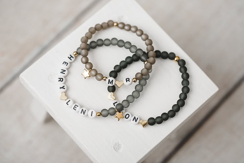 May include: Three beaded bracelets with the names "Henry", "Leni", and "Leon" spelled out in white beads. Each bracelet has a gold star and heart charm.