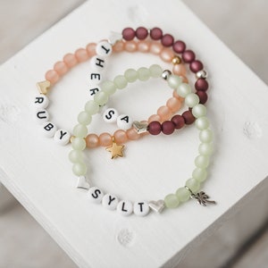 May include: Three beaded bracelets with name charms. The top bracelet is burgundy, the middle bracelet is peach, and the bottom bracelet is light green. The charms spell out the names "Ruby", "Theresa", and "Sylt".