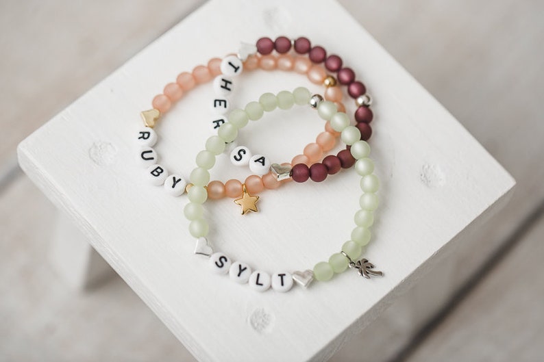 May include: Three beaded bracelets with name charms. The top bracelet is burgundy, the middle bracelet is peach, and the bottom bracelet is light green. The charms spell out the names "Ruby", "Theresa", and "Sylt".