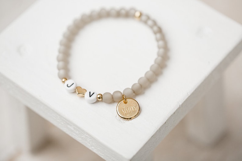 May include: A gray beaded bracelet with white letter beads spelling "JV" and a gold heart charm. The bracelet has a gold charm with the word "Sisters" engraved on it.