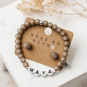 Könnte beinhalten: Ein braunes Perlenarmband mit einem silbernen Sternanhänger und dem Namen "MAYA" in weißen Perlen. Das Armband wird auf einer braunen Karte mit dem Text "WEAR ME WITH" und einem kleinen rosa Herz präsentiert.