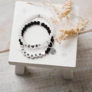 May include: Two beaded bracelets with black and white beads. One bracelet has the name "Torben" spelled out with white beads and a silver heart charm. The other bracelet has the letters "CB" spelled out with white beads and a silver heart charm with a tree design.