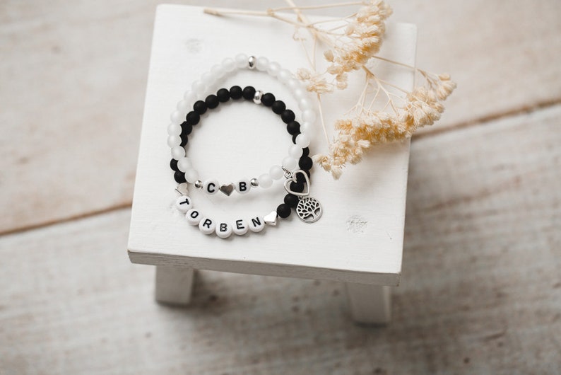 May include: Two beaded bracelets with black and white beads. One bracelet has the name "Torben" spelled out with white beads and a silver heart charm. The other bracelet has the letters "CB" spelled out with white beads and a silver heart charm with a tree design.