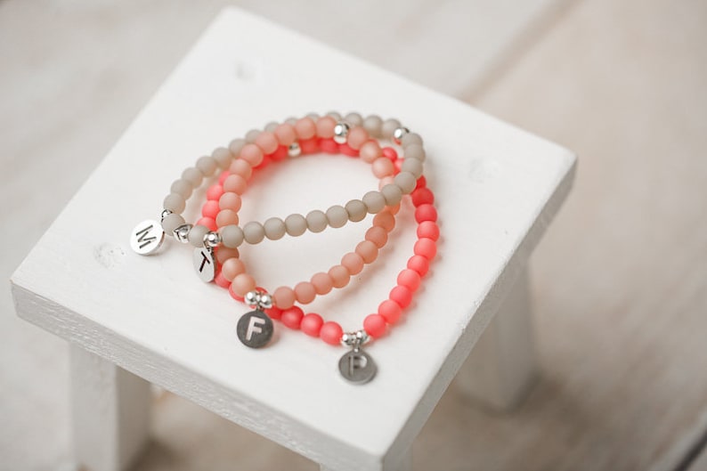 May include: Three beaded bracelets with silver charms. The bracelets are layered, with the top bracelet being a light pink color, the middle bracelet being a light beige color, and the bottom bracelet being a coral color. The charms are round and have the letters "M", "F", and "T" on them.