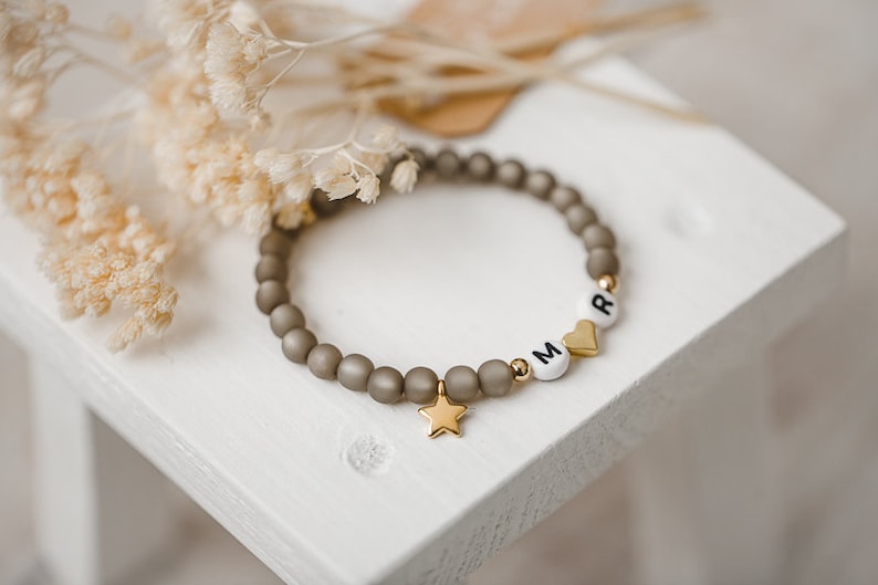 May include: A gray beaded bracelet with gold accents and white letter beads spelling "MR" with a gold heart charm and a gold star charm.