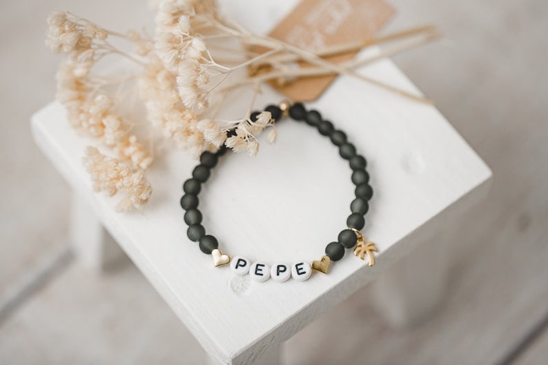 May include: A black beaded bracelet with a gold palm tree charm and white letter beads spelling "PEPE".
