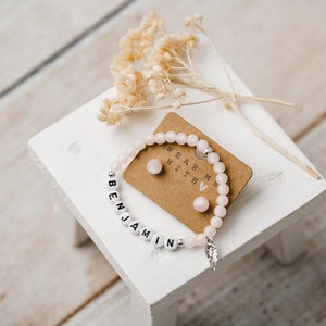 May include: A pink beaded bracelet with the name "Benjamin" spelled out in white beads. The bracelet has a small silver feather charm. The image also shows a small card with the text "Wear me with" and two pink heart-shaped earrings.