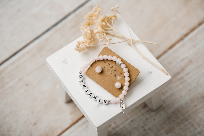 May include: A pink beaded bracelet with the name "Benjamin" spelled out in white beads. The bracelet has a small silver feather charm. The image also shows a small card with the text "Wear me with" and two pink heart-shaped earrings.