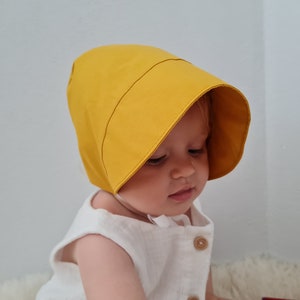 Könnte beinhalten: Ein Baby trägt einen gelben Sonnenhut mit einem weißen Kleid.