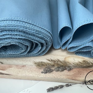 Puede incluir: Un rollo de tela azul claro con un borde cosido. La tela está doblada y enrollada, y los bordes son visibles. La tela está sobre una superficie de madera.