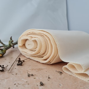 Op de afbeelding: Een rol crèmekleurige stof met een overlockrand. De stof is strak opgerold, met het uiteinde van de rol zichtbaar. De stof ligt op een lichtbruin oppervlak, met een kleine tak en gedroogde bloemen op de achtergrond.