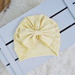 Könnte beinhalten: Ein blassgelber Turban mit einer großen Schleife oben. Der Turban ist aus einem weichen, dehnbaren Stoff.
