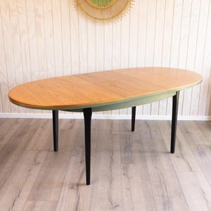 May include: An oval wooden dining table with a light brown tabletop and a green border. The table has four black legs. The table is approximately 152 cm long and 91 cm wide.