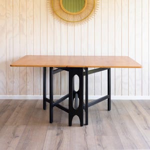May include: A rectangular wooden table with a light brown top and black legs. The table is a drop-leaf design. The table is approximately 122 cm long and 76 cm wide when fully extended.