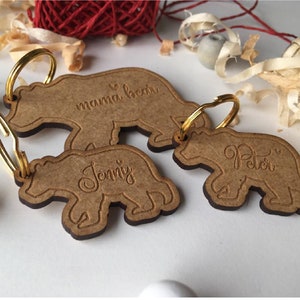 Può includere: Tre portachiavi in legno a forma di orso con anelli dorati. Il portachiavi superiore dice "mama bear", quello centrale dice "Jenny" e quello inferiore dice "Peter".