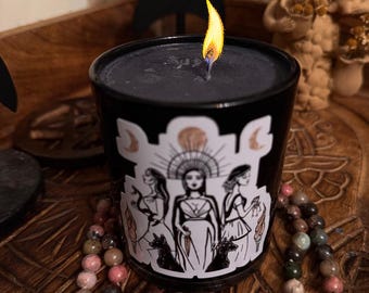May include: A black candle with a white sticker featuring a drawing of three women with a sun and moon design. The candle is lit and sitting on a wooden surface with a beaded necklace.
