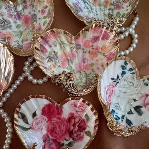 May include: Heart-shaped decorative seashell dishes with floral designs in pink, red, and purple. Each dish has a gold-colored ornate base and rim. A pearl necklace is draped across some of the dishes.