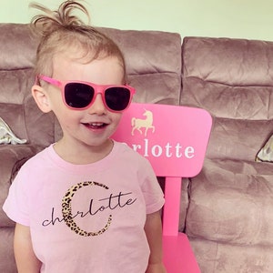 May include: A pink child's chair with a unicorn design and the name "Charlotte" written on it. A young child wearing pink sunglasses and a pink shirt with a leopard print "C" and the name "Charlotte" is sitting in the chair.