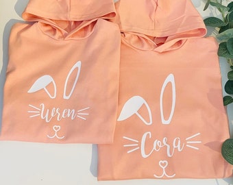 May include: Two peach-colored hooded sweatshirts with white bunny ears and the names "Wren" and "Cora" printed on the front.