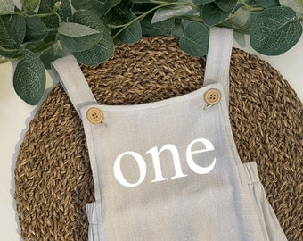 May include: A gray linen baby onesie with the word "one" printed in white on the front. The onesie has two brown buttons on the straps.