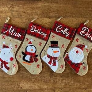 May include: Four personalized Christmas stockings with red and white plaid trim. The stockings feature embroidered names: Mum, Bethany, Colby, and Dad. Each stocking has a different festive character: a reindeer, a penguin, a snowman, and Santa Claus.