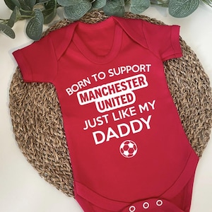 May include: Red baby bodysuit with white text that reads "BORN TO SUPPORT MANCHESTER UNITED JUST LIKE MY DADDY" and a soccer ball graphic. The bodysuit has short sleeves and three snaps at the bottom.