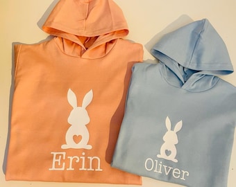 May include: Two hooded sweatshirts with bunny silhouettes and names "Erin" and "Oliver" printed on them. The sweatshirt on the left is peach colored and the sweatshirt on the right is light blue.