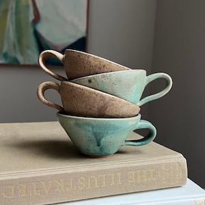 May include: A stack of four ceramic teacups in shades of brown and teal, each with a curved handle. The cups are stacked on top of two books with gold lettering. The background includes a painting and a neutral wall.