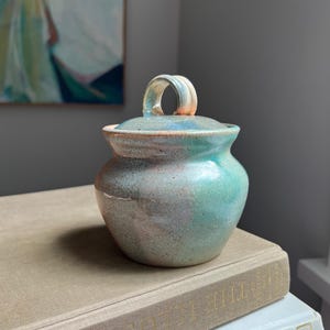 May include: A small, round, handmade ceramic jar with a lid featuring a looped handle. The glaze displays a mottled blend of soft pastel colors, including shades of mint green, pale blue, and muted pink, with a smooth, glossy finish throughout.