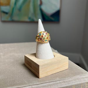 May include: A gold-toned ring with a dome shape, encrusted with a multitude of colorful gemstones. The ring is displayed on a white cone-shaped stand set within a small, light-colored wooden base. The ring's design features a cluster of various colored gems.