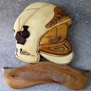 Peut inclure: Une plaque murale en bois représentant un casque en bois sculpté avec un motif brun et noir. Le casque est monté sur une base en bois incurvée. Le texte "MSA CALLST" est gravé dans le casque.
