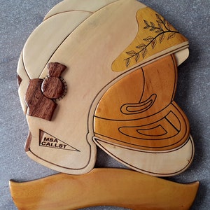 Puede incluir: Una decoración de pared de madera con forma de casco de bombero con el texto "MSA CALLST" en el lateral. El casco está hecho de madera clara y oscura y tiene un diseño decorativo de hojas.