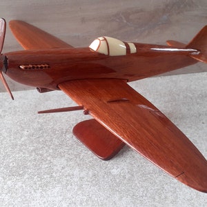 Puede incluir: Maqueta de avión de madera con acabado marrón rojizo oscuro. El modelo presenta una hélice, alas y una cabina con capota color crema. El avión está montado sobre un soporte de madera a juego.