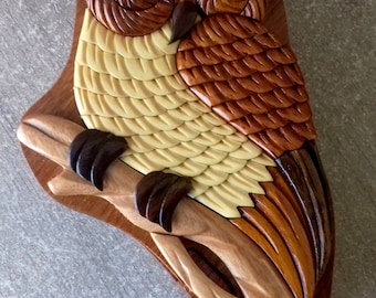 Op de afbeelding: Een houten puzzeldoos in de vorm van een uil met ingewikkelde details. De uil zit op een tak en heeft een bruin, geel en oranje kleurenschema.