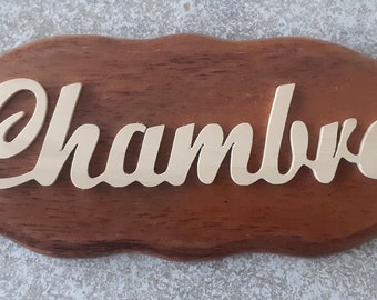 May include: A wooden sign with the word "Chambre" in gold lettering. The sign is oval shaped and has a rustic brown finish.
