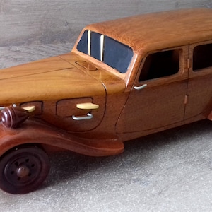 May include: A wooden model car with a brown finish. The car has a detailed front grille and headlights. The car is a vintage style sedan.