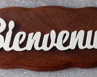 May include: A wooden sign with a carved welcome message in white lettering. The sign is shaped like an oval and has a dark brown wood grain.