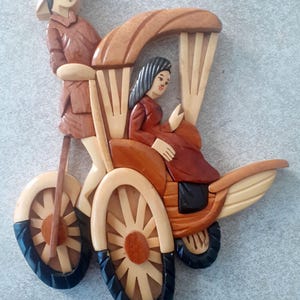 Może przedstawiać: Drewniana rzeźba tradycyjnego wietnamskiego cyclo, czyli trójkołowego taksówki rowerowej, z dwiema postaciami. Cyclo jest pomalowane w odcieniach brązu i czerni, a postacie ubrane są w tradycyjne stroje. Rzeźba jest popularnym pamiątkowym przedmiotem.